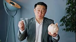 Laptop web camera view of confident japanese male holding beige piggy bank in one hand and metal coin in other in office environment. Man with glasses telling about benefits of savings indoors.
