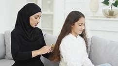 Young beautiful Arabian woman in black hijab smiling and combing long hair of pretty small teen girl with comb. At home on couch. Muslim mother making hair style for little teenage daughter in room.