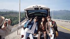 4K Happy Asian man and woman enjoy and fun outdoor lifestyle road trip countryside travel on summer holiday vacation. Generation z people friends sit on car trunk using camera taking picture together.