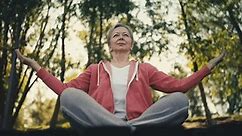 Calm senior woman doing breathing exercise on lake shore, sitting in lotus pose