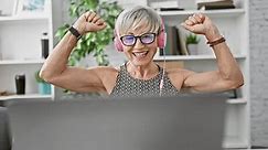 A joyful mature woman with grey hair wearing headphones celebrates at her office desk, embodying positivity and success indoors.