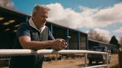 Mature male farm worker standing outside barn leaning on fence looking at mobile phone - shot in slow motion