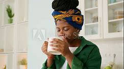 African American girl with braids and colorful headband is staying in kitchen, drinking tea. Dark skinned young woman takes sip, savoring taste and enjoying it. She feels peaceful and happy.