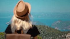 Girl with backpack and hat on top of mountain looking from above at beautiful mountain valley with sea or ocean and islands. Landscape with sporty young blonde woman sitting on rock. Hiking. Nature