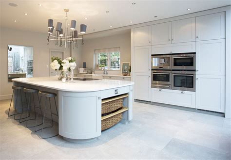 A white kitchen island is fitted with a marble countertop and a set of low back wicker island stools under four white drum light pendants. Shaker style kitchen with a curved island unit featuring ...