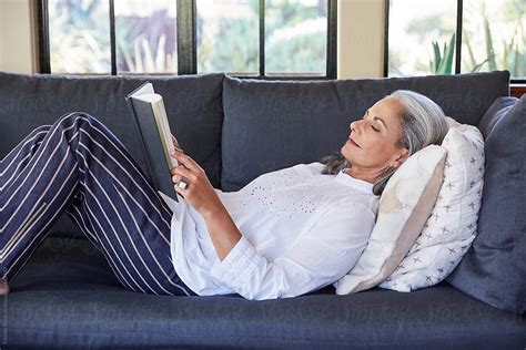 Mature Woman With Grey Hair Reading A Book Lying Down On Sofa In Living ...